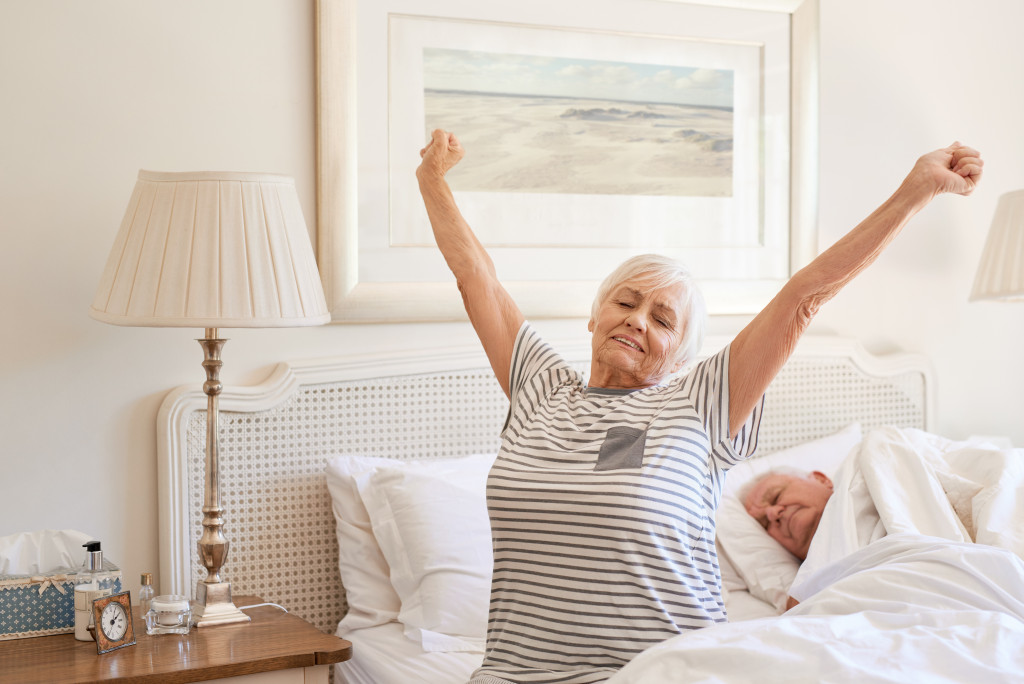 Senior woman stretching hers arms in the morning while sitting on her bed with her husband still sleeping behind her.